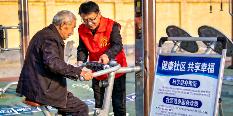 社区志愿者在指导居民使用健身器材，旁边有政策宣传展板